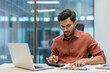 © Liubomir - Young male entrepreneur working in office using calculator and laptop. Serious and focused on financial calculations, seated indoors at modern workplace. Represents business and productivity