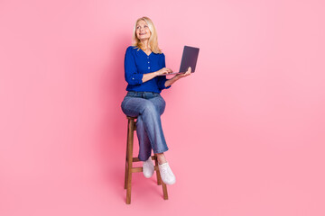 Wall Mural - Full length photo of lovely pensioner lady sit chair hold netbook look empty space dressed blue garment isolated on pink color background