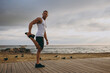 © ViDi Studio - Young strong sporty toned fit sportsman man wear sports clothes earphones raise up leg holding foot warm up training at sunrise sun dawn over sea beach outdoor on pier seaside in summer day morning