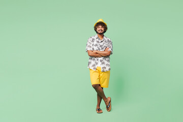 Poster - Full body young smiling happy Indian man wear beach shirt hat rest near hotel pool hold hands crossed folded look camera isolated on plain green color background. Summer vacation sea sun tan concept.