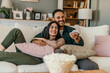 © La Famiglia - Diverse couple, watching tv and remote on living room couch with smile, hug or relax with click for choice, show or film