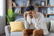 © Tetiana - Stressed man sitting on sofa holding head in hands feeling overwhelmed in modern living room environment