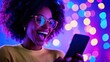 © Anna - Excited woman with glasses using smartphone against colorful Bokeh lights. Online shopping excitement during Cyber Monday.