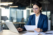 © Liubomir - Asian businesswoman in professional suit examining documents while using laptop in modern office. Image conveys focus, dedication, and productivity with digital technology and paperwork.