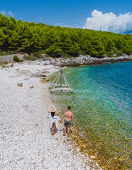 Wall Mural - A couple leisurely walks hand in hand along the pebbled beach of Brac Island, surrounded by lush greenery and the sparkling Adriatic Sea under a bright blue sky, enjoying a peaceful summer moment.