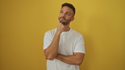 Poster - Thinking young hispanic man wearing white t-shirt, standing, finger on chin, looking up, pondering doubt over isolated yellow background, concentrated and wondering thoughtfully