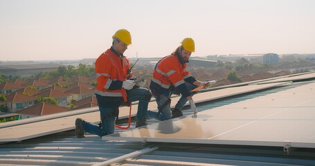 Two engineers solar technicians in reflective gear discussing working examine solar installations on rooftop panels at dusk, with suburban backdrop