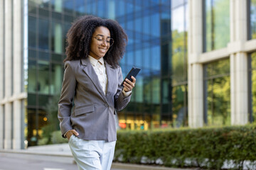 Confident businesswoman in formal attire walking outside modern office building using smartphone. Woman appears engaged and professional, enjoying sunny day.