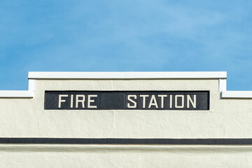 A white vintage concrete building with a black band. The retro fire station's exterior wall has a black background with white lettering indicating a fire station. The background is a pale blue sky.