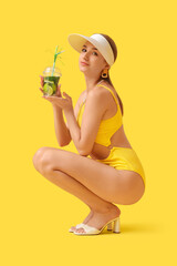 Wall Mural - Beautiful young woman in swimsuit with cup of mojito sitting on yellow background