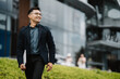 © Kritdanai - Confident Stride: A young Asian businessman exudes confidence and success as he walks towards his goals with a laptop in hand. The cityscape backdrop adds a sense of ambition and progress.