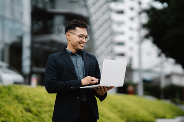 Urban Professional in Action: Young Asian businessman in glasses smiles at his laptop, navigating the city with confidence and efficiency.