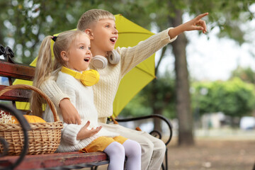 Poster - Children with headphones and yellow umbrella on bench pointing at something in park