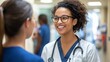 © Intania - A Smiling Female Doctor Wearing Glasses and a Stethoscope, Talking to Another Person in a Hospital Corridor