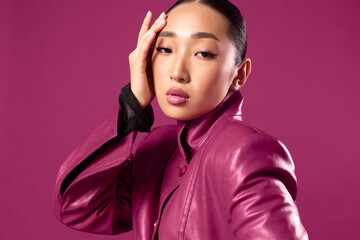 Wall Mural - Fashion asian brunette in purple leather jacket and black shirt posing against purple background with hands on head, looking at camera