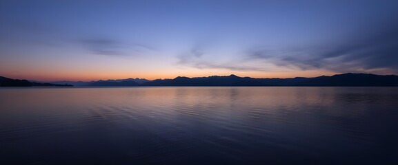 Poster - A landscape shot at dusk by the edge of a lake. Mountains on the horizon and the slightly blurred sky reflect on the calm surface of the water.