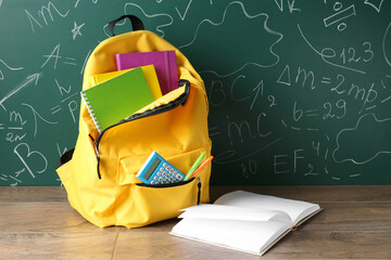 Sticker - Backpack with different school stationery on wooden table against green chalkboard