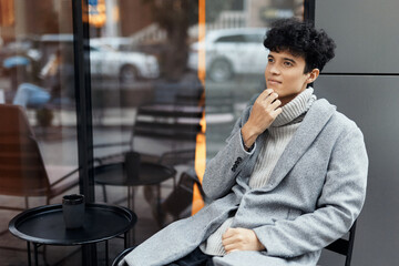 Fashionable young man in a gray coat sitting by a window holding a cup of coffee