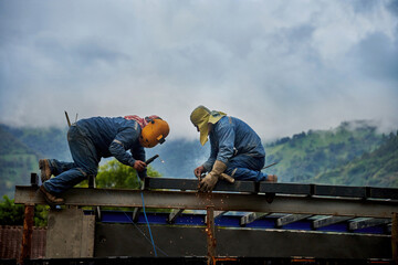 two construction workers welding steel beams on a building structure, with a mountainous landscape i