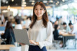 © Ganesha - Smiling Japanese businesswoman standing in an office, holding her laptop. She is wearing casual clothes, and other people working behind her are blurred.,generated ai.