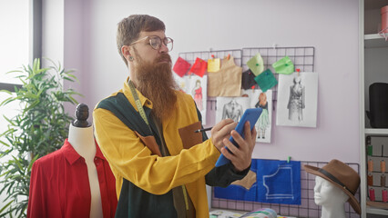 Poster - Bearded man using smartphone in tailor shop with mannequin and fashion sketches.