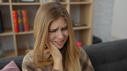 Poster - A distressed young woman with blonde hair feels toothache in a cozy living room setting.