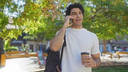 Poster - Handsome young hispanic man in an urban park enjoying a coffee while talking on his phone outdoors