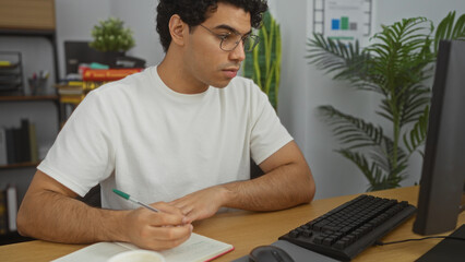 Poster - Hispanic man working in an office setting, focusing on a notebook while using a desktop computer, surrounded by indoor plants and shelves.