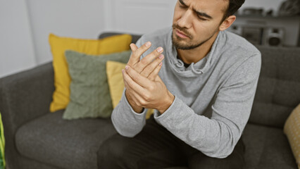 A hispanic man examines his hand in a modern living room, showing potential pain or injury concern.