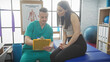 © Krakenimages.com - A man in teal scrubs attentively reviews notes on a clipboard as a woman in black sits beside him in a well-equipped physiotherapy clinic.