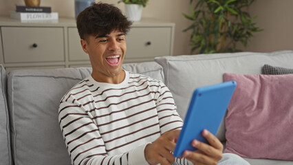 A young, hispanic man is smiling and looking at a tablet while sitting on a comfortable sofa in his home living room, surrounded by casual decor and greenery.