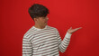 © Krakenimages.com - A handsome young hispanic man in a striped shirt presents something with his hand extended against a bold red background.