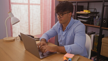 Wall Mural - Young hispanic man working on a laptop in a modern office setting with bookshelves and organized workspace.