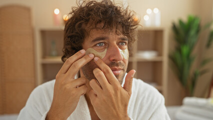 Poster - Young man applying eye patches in a spa wellness center wearing a white bathrobe in an indoor beauty salon