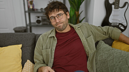 A casual hispanic man with a beard and glasses lounges on a sofa at home, surrounded by cushions and a guitar in the background.