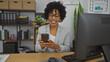 © Krakenimages.com - Young african american woman with curly hair smiling while using a phone in an office interior with bookshelves and plants.
