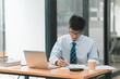 © phaitoon - A focused professional writes notes at a desk, using a laptop amidst a modern office environment.