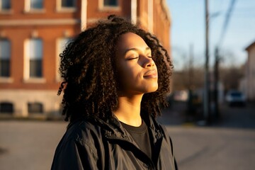 A person with curly hair stands outdoors with eyes closed, enjoying the sunlight, wearing a dark jacket. Background shows a building and a street.