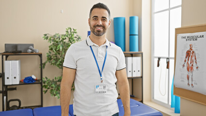 Canvas Print - A smiling hispanic man with a beard stands confidently in a clinic, portraying a professional healthcare atmosphere.