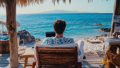Poster - A man is sitting on the beach with his laptop, working remotely, enjoying the beautiful sea view. He has short hair and wears a blue printed shirt. The sun shines brightly over the clear water