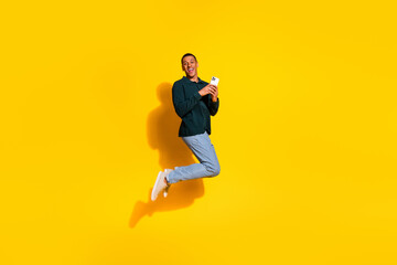 Wall Mural - Full size photo of handsome young guy jump hold gadget dressed stylish green garment isolated on yellow color background