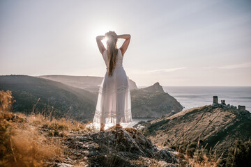 Wall Mural - A woman stands on a hill overlooking the ocean. She is wearing a white dress and has her hands on her head. The scene is serene and peaceful, with the woman looking out over the water.