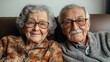 © Nataliia_Trushchenko - Two elderly people are sitting on a couch, smiling at the camera. The man is wearing glasses and the woman has a scarf around her neck. Scene is warm and friendly