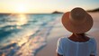 ©  lukaPixMedia - An image of a person with a straw sunhat, viewed from the back, standing on a tranquil beach as they watch the serene sunset, reflecting on the gentle waves of the ocean.