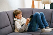 © Irina Schmidt - Teenager boy lounging comfortably on a grey couch while using a tablet. Child is relaxed, holding the tablet with both hands, and appears to be engaged in what he is watching or reading, or gaming