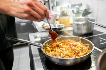 Wall Mural - A close-up of a hand stirring cooked food with tomato sauce in a pan, capturing the action of cooking. The vibrant red sauce contrasts with food, highlighting texture and freshness of the ingredients.