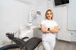 © Vadim - A woman stands in front of a dental chair in a dental office. She is wearing a white shirt and white pants