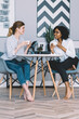 © BullRun - Positive dark skinned young woman communicating with cheerful caucasian friend and discussing last news from fashion world during warm conversation in modern apartment with stylish design interior