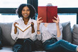© BullRun - Portrait of happy smiling afro american female looking at camera while sitting barefoot on comfortable couch near friend reading interesting literature bestseller in apartment with stylish interior