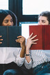 © BullRun - Multicultural female readers looking each other in eyes while sitting on comfortable couch and holding literature books near faces having joyful mood for weekend, concept of knowledge and education
